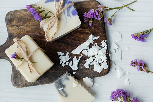 Top view of homemade soap near flakes on cutting board and flowers on table