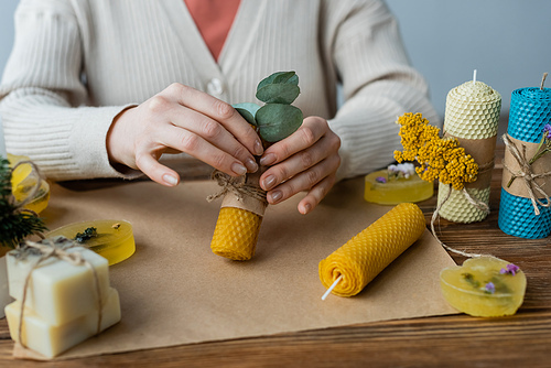 Cropped view of craftswoman decorating handmade candle with eucalyptus at home