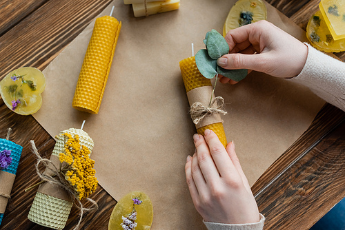 Top view of craftswoman decorating handmade candle with eucalyptus near soap bars at home