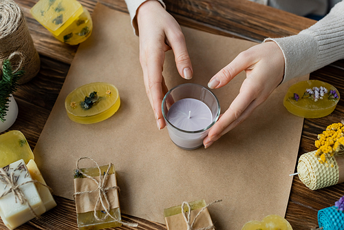 Top view of craftswoman holding handmade candle in glass near soap bars on craft paper