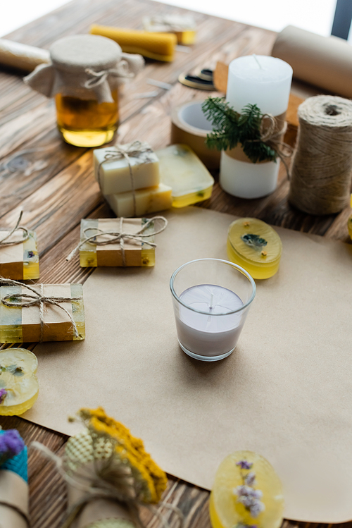 Handmade candle in glass near soap bars and craft paper on wooden surface