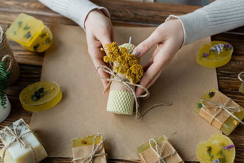 Top view of craftswoman holding handmade candle with flower near soap bars on craft paper