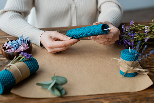 Cropped view of blurred craftswoman making candle with wick near flowers on table