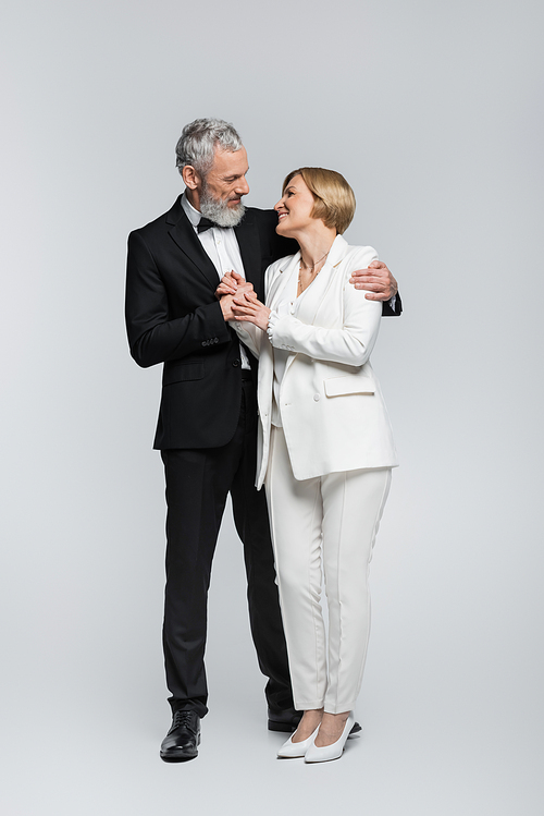 Full length of grey haired groom hugging bride in suit on grey background