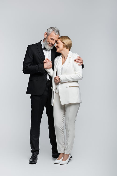 Positive mature bride in suit holding hand of groom on grey background
