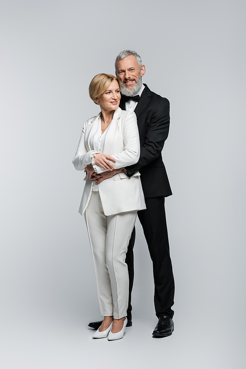 Positive groom hugging mature bride in suit on grey background