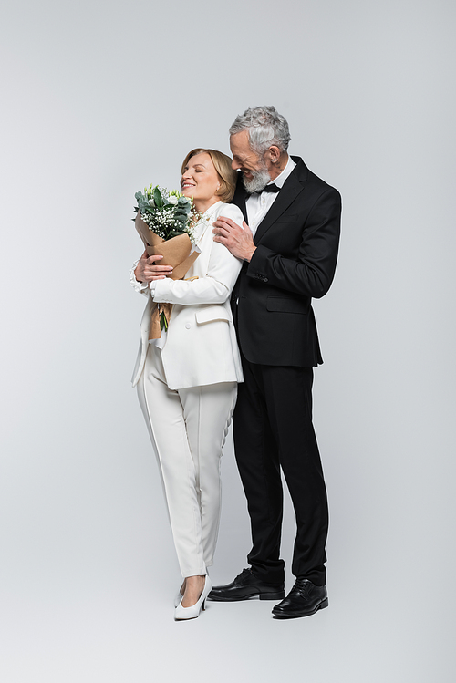 Mature groom hugging cheerful bride with bouquet on grey background