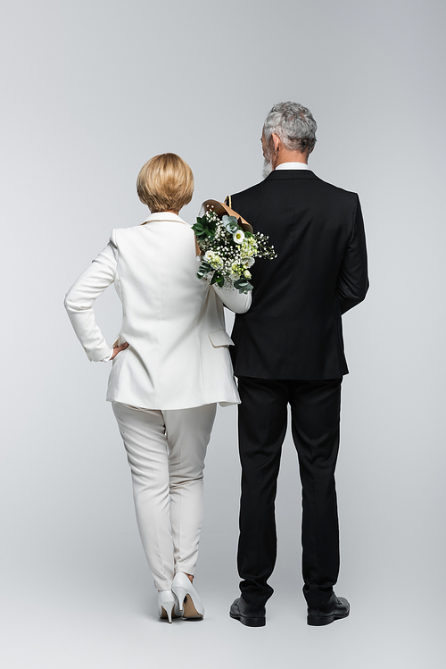 Back view of bride in suit holding wedding bouquet near groom on grey background