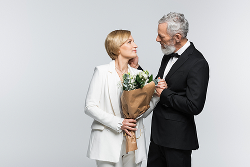 Side view of mature groom touching bride with wedding bouquet isolated on grey