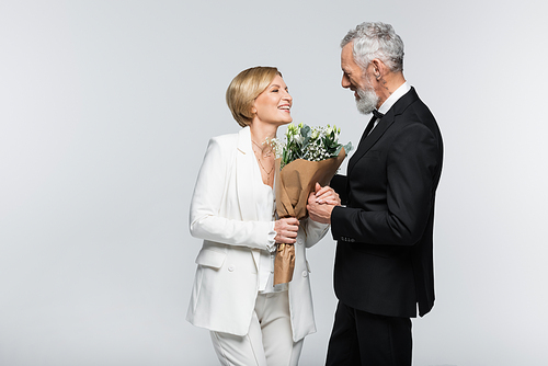 Positive mature bride holding bouquet and hand of groom isolated on grey