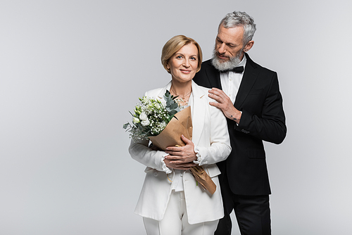 Mature groom in suit hugging smiling bride with wedding bouquet isolated on grey
