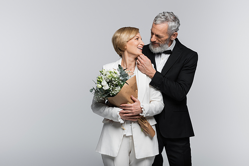 Mature groom touching chin of smiling bride with bouquet isolated on grey