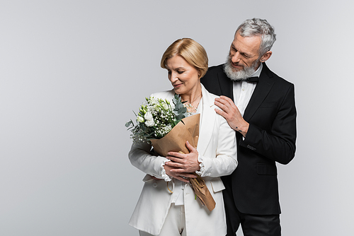 Positive groom in suit hugging middle aged bride with bouquet isolated on grey
