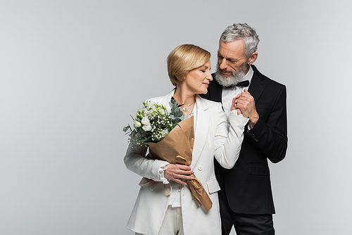 Middle aged groom holding hand of bride with wedding bouquet isolated on grey