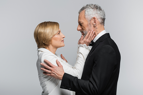 Side view of mature bride touching face of groom in suit isolated on grey
