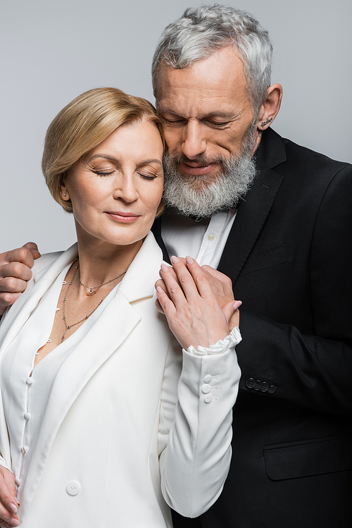 Portrait of bearded groom with closed eyes hugging bride in suit isolated on grey