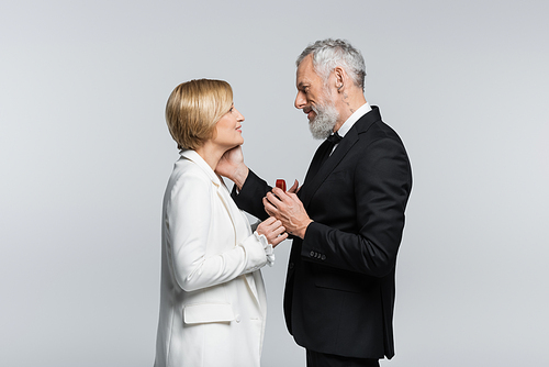Side view of mature man holding jewelry box and touching smiling woman isolated on grey