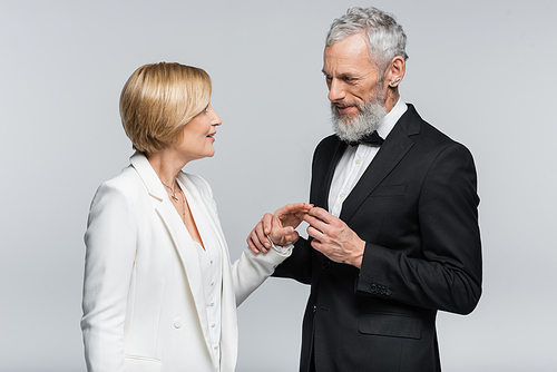 Middle aged groom wearing wedding ring on smiling bride isolated on grey