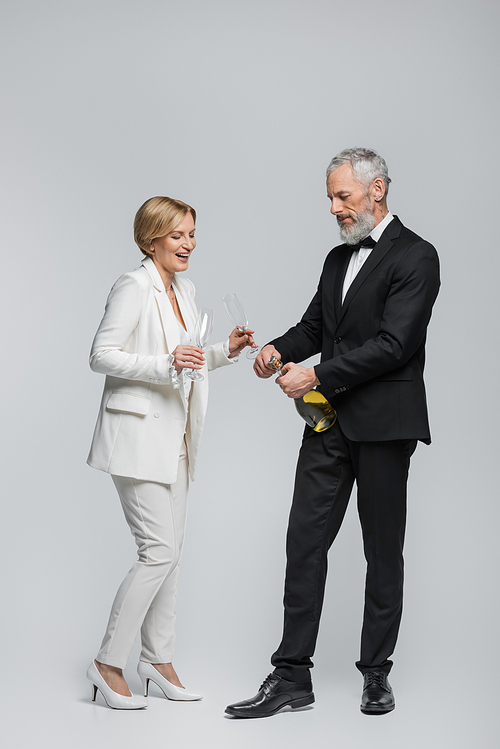 Cheerful mature bride holding glasses near groom with bottle of champagne on grey background
