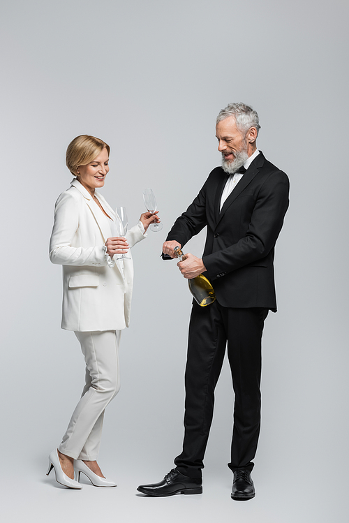 Positive middle aged bride holding glasses near groom with champagne on grey background