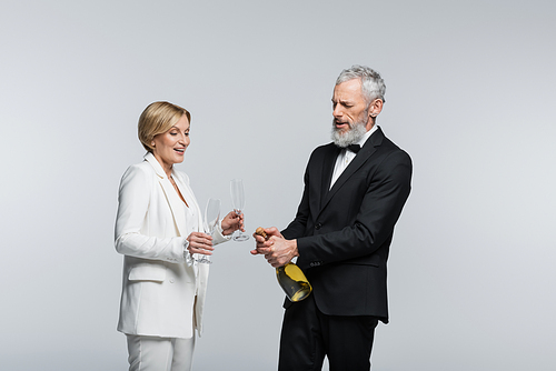 Mature groom opening champagne near positive bride with glasses isolated on grey
