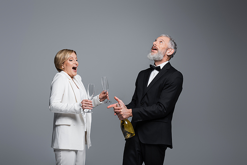 Excited mature groom opening champagne near bride with glasses isolated on grey