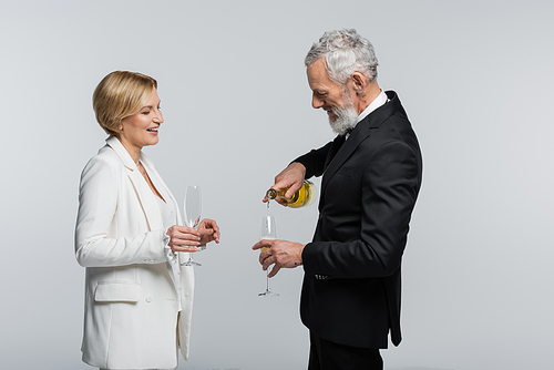 Smiling mature groom pouring champagne near bride with glass isolated on grey