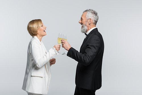 Side view of positive bride clinking champagne with mature groom isolated on grey