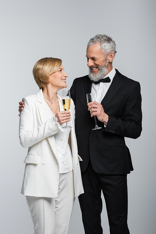 Positive mature groom hugging bride with glass of champagne isolated on grey
