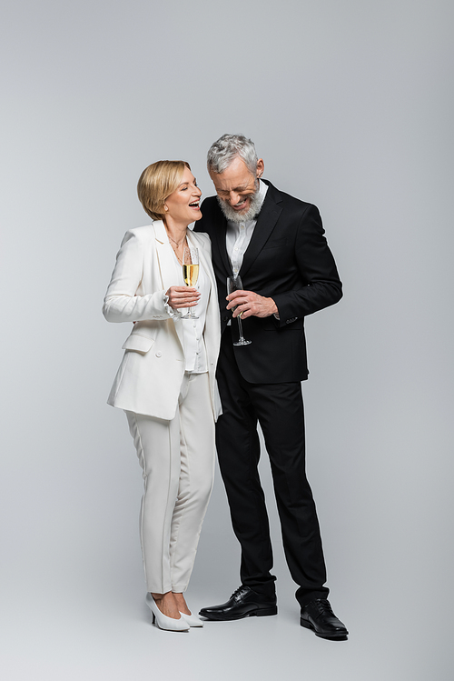 Happy mature bride holding glass of champagne near groom in suit on grey background
