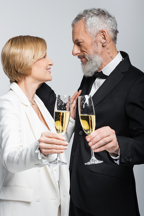 Side view of happy mature woman holding glass of champagne near groom isolated on grey