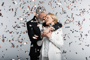 Mature groom kissing and hugging bride under confetti on grey background