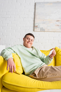 Positive teenager with down syndrome holding smartphone while sitting on couch at home