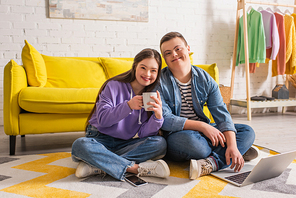 Positive couple of teenagers with down syndrome holding cup and using laptop at home