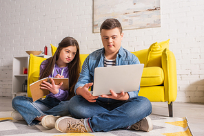 Teenager with down syndrome writing on notebook near friend with laptop at home