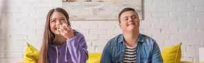 Smiling teenager with down syndrome holding popcorn near friend on couch at home, banner