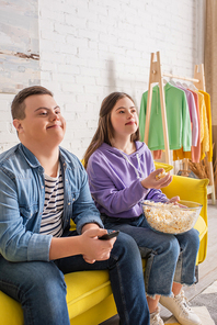 Positive boy with down syndrome holding remote controller near friend with popcorn at home