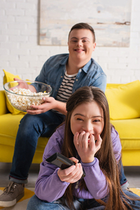 Positive girl with down syndrome holding remote controller near blurred friend with popcorn at home