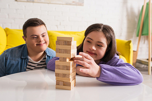 Smiling teen girl with down syndrome playing wood blocks game near friend at home