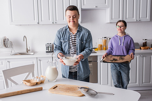 Teenager with down syndrome holding flour near girlfriend with baking sheet in kitchen