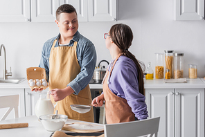 Smiling teenager with down syndrome holding ingredients near girlfriend in apron in kitchen