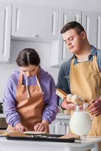 Girl with down syndrome cooking near friend with eggs and baking sheet in kitchen