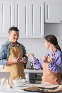 Positive teenager with down syndrome holding smartphone near friend with dough in kitchen