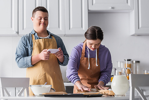 Teenager with down syndrome making dough near ingredients and friend holding smartphone in kitchen