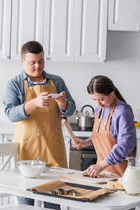 Teenager with down syndrome cooking dough near friend with smartphone at home