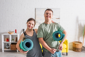 Positive teen couple with down syndrome holding fitness mats at home