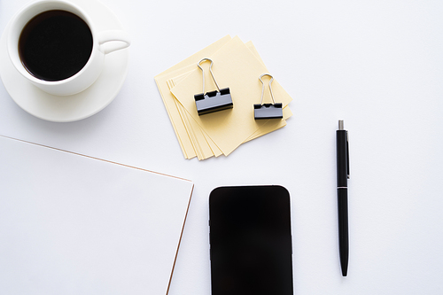 top view of mobile phone with blank screen near cup of coffee and stationery on white