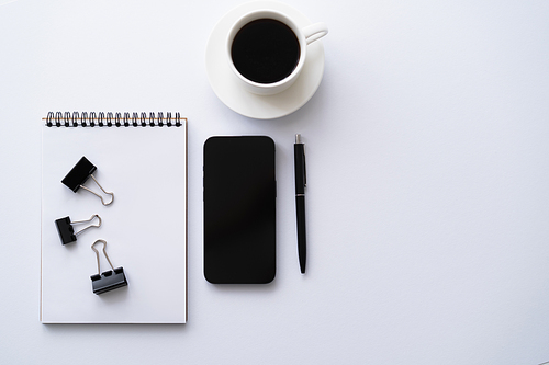 top view of cellphone with blank screen near cup of coffee and stationery on white