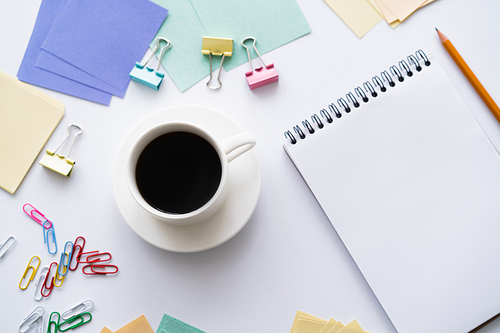top view of cup of coffee near blank notebook and colorful stationery on white