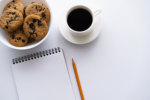 top view of bowl with cookies with chocolate chip near cup of coffee and notebook on white